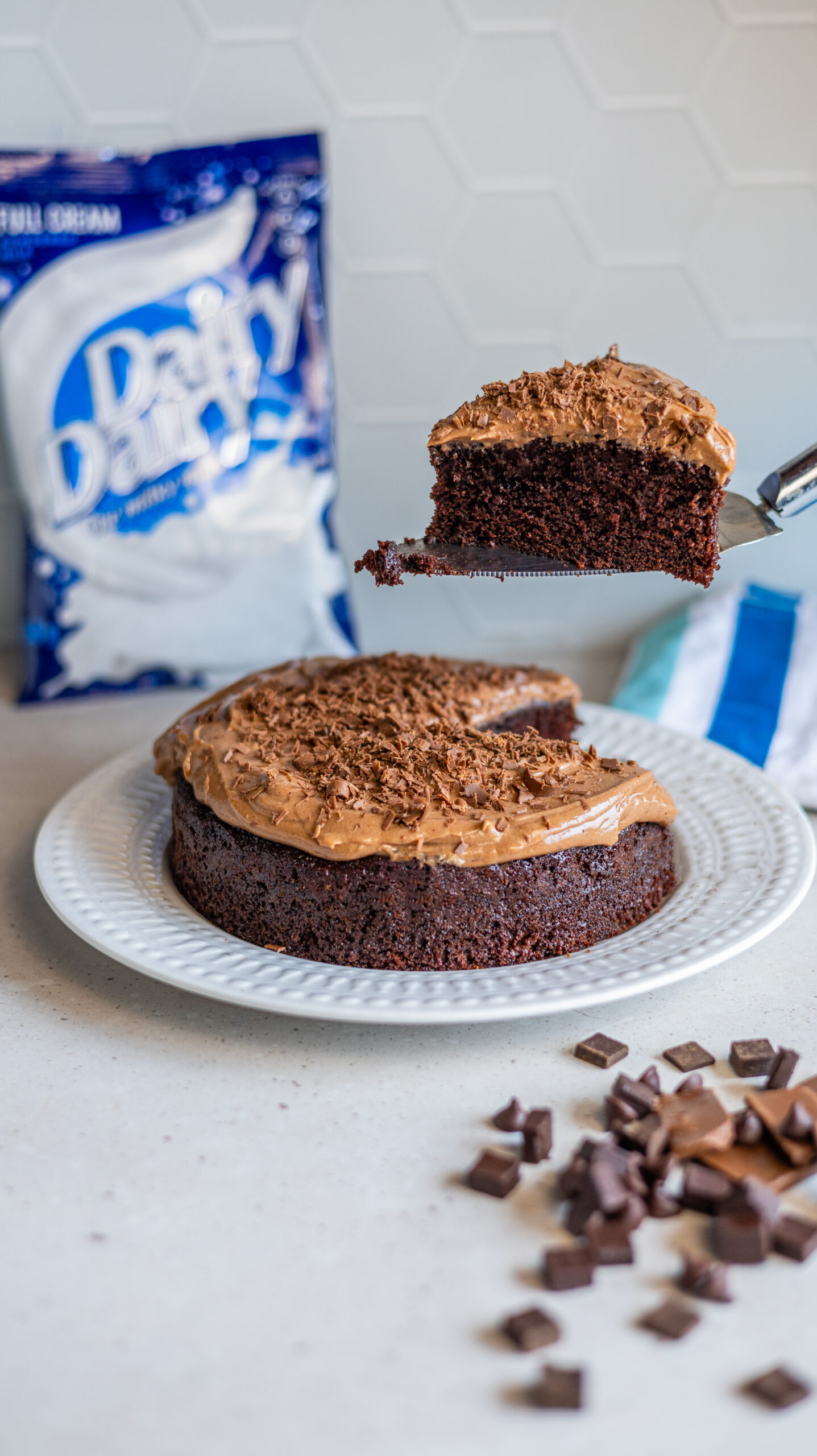 8-inch round chocolate cake with chocolate buttercream icing on top, sprinkled with milk chocolate shavings. A slice has been cut from the cake and is being held aloft just above the cake itself on a spatula. A pack of Dairy Dairy Full Cream Milk is in the background to the left. Presented as a Father's Day treat.